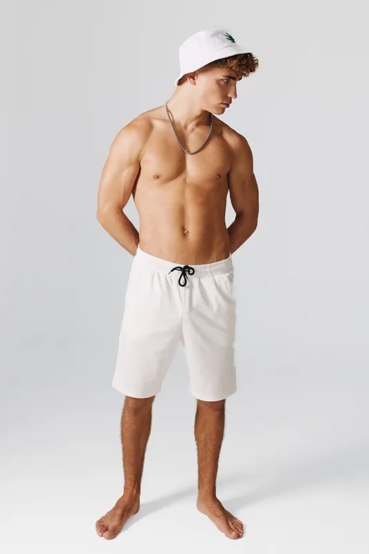 Young man with curly hair wearing white bucket hat and white shorts stands barefoot against empty background, looking to the side with hands behind back. Fitness and casual summer style.