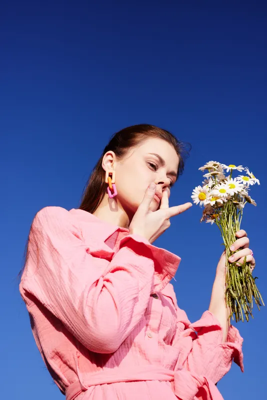 Young woman in pink dress holding daisies against a clear blue sky, expressing joy and connection with nature, vibrant seasonal look