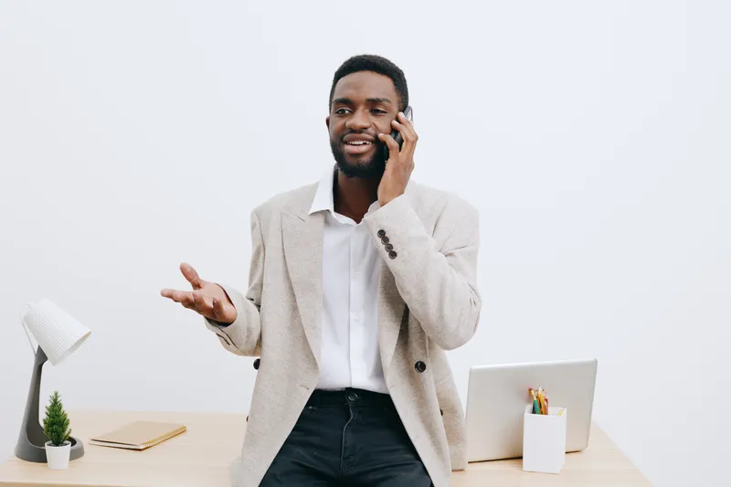 Friendly smiling man talking on mobile phone in modern office interior with white background, business casual attire, relaxed and confident mood, using technology for communication and work. People co