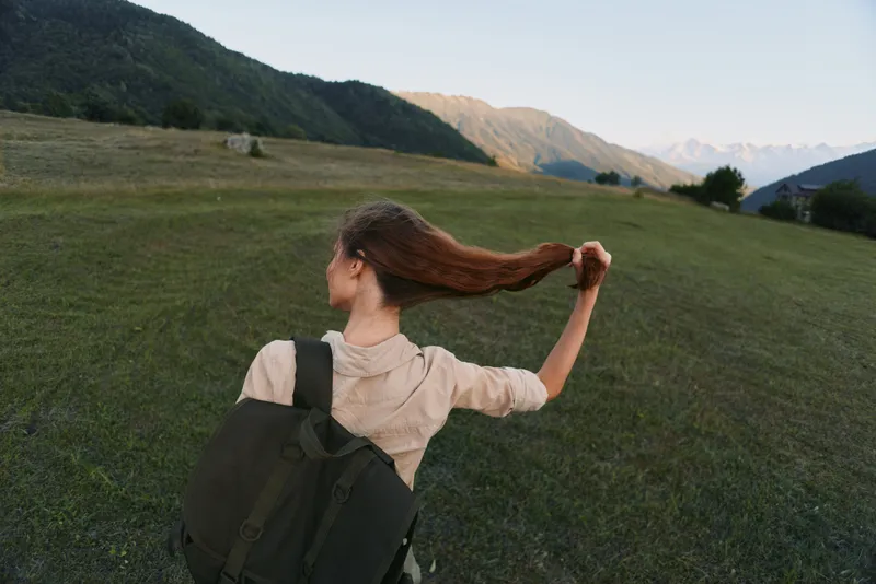 Young woman with backpack walks across a grassy meadow, hair swinging behind, toward distant mountains, expressing calm and freedom.