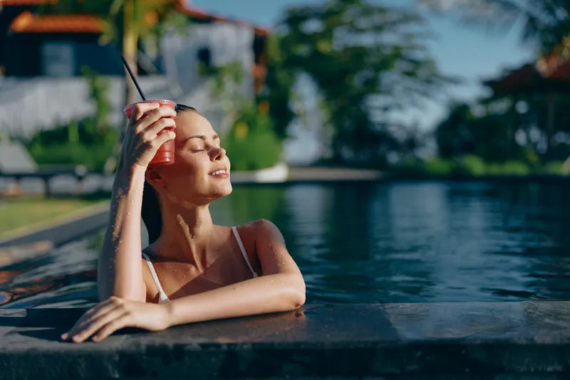 A young woman relaxes at poolside, leaning on the edge and enjoying sun, wearing a white swimsuit and sunglasses, smiling softly in a carefree moment.