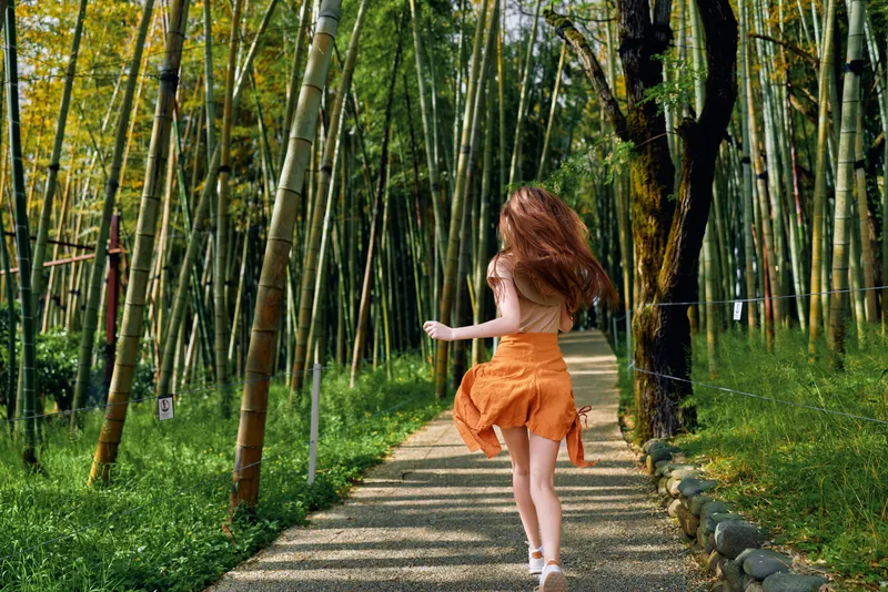 Woman girl running on a stone path through a bamboo forest wearing an orange dress, summer scene with motion and sunlight filtering through green stalks, carefree outdoor activity