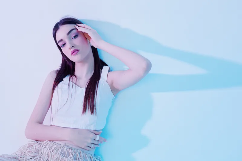 Young woman with long dark hair poses in a white sleeveless top against a pastel background. Soft lighting highlights her beauty and fashion style in a studio setting.
