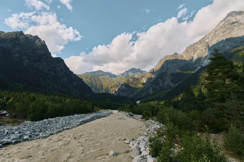 A serene landscape shows a rocky riverbed winding through a valley flanked by towering mountains under a bright sky. Lush trees and calm air create a peaceful outdoor scene.