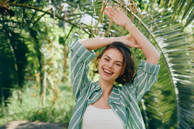 Joyful woman outdoors, smiling and dancing with arms raised, wearing a striped shirt; lush greenery and sunlight create a lively, carefree mood.