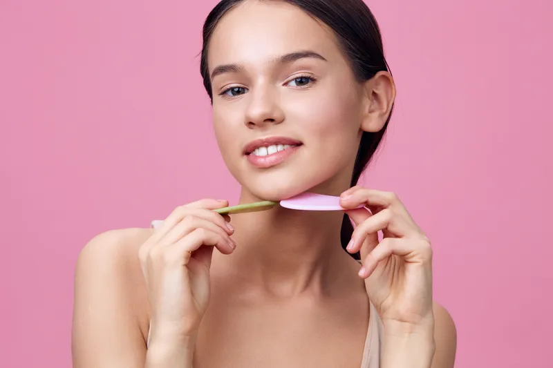 Young woman smiling while holding a pink and green facial tool against a pink background, showcasing skincare and beauty trends