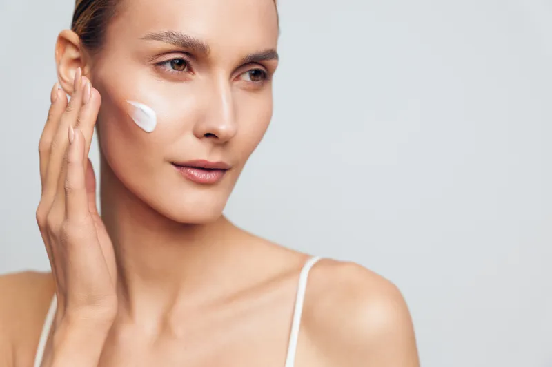 Young woman applying white face cream on cheek with fingers. Close-up portrait on light background. Concept of skincare, beauty, and daily self-care routine with soft natural lighting.