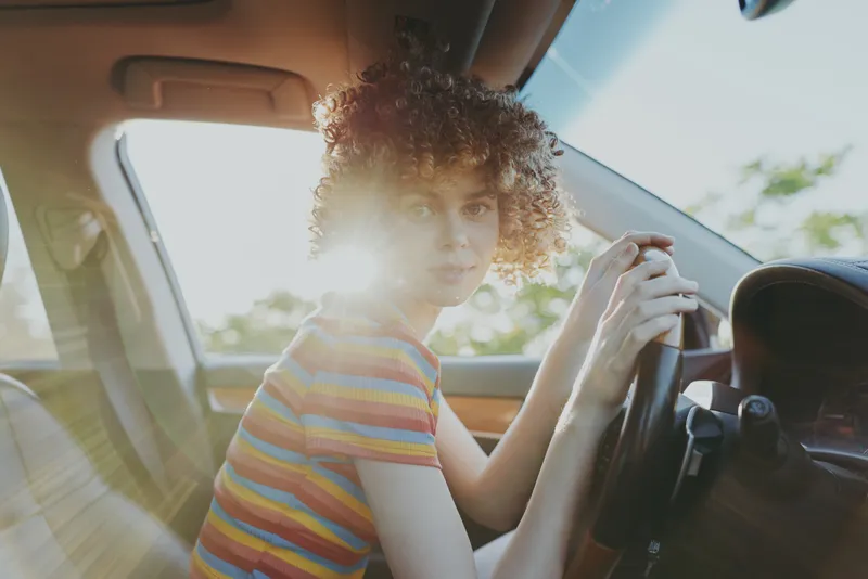 Casual portrait of a young girl in a colorful striped shirt sitting in a car, holding the steering wheel, bathed in warm sunlight, conveying a relaxed lifestyle and retro film color