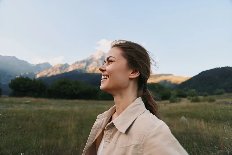 Woman lifestyle portrait outdoors in open fields with mountains, smiling and relaxed, wearing light jacket, enjoying natural scenery and warm sunlight.