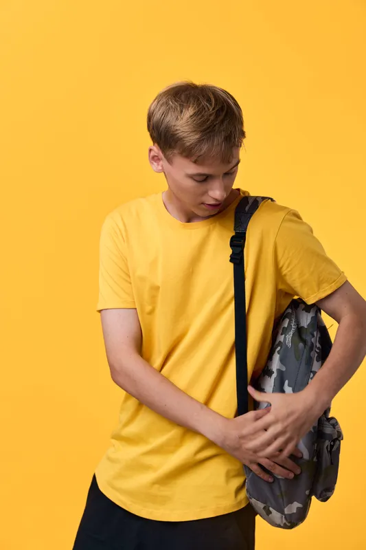 Boy in a yellow t shirt with a backpack adjusting the strap against a bright yellow background, casual school style, ready for class, travel, or daily routine