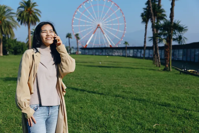 Woman in an outdoor park with a distant ferris wheel, wearing a beige coat and sweater, enjoying sunny leisure and a candid moment outside during spring