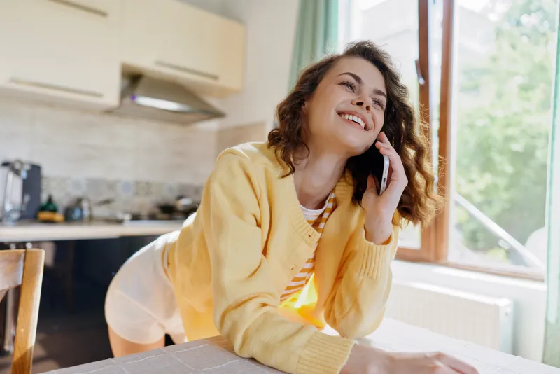 Casual woman at home speaks on phone in sunny kitchen, wearing a yellow cardigan, relaxed smile, natural light, everyday lifestyle, candid moment of domestic life