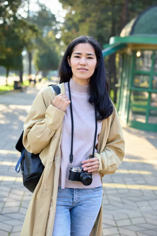 Casual woman outdoors with camera and backpack in a park, beige coat and sweater, natural light, relaxed mood, street style photography in nature