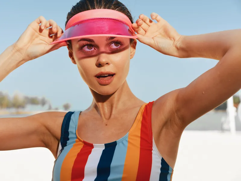 A young woman in a bright striped swimsuit adjusts a pink visor at the sunny beach, her expression confident as azure sky and soft sand create a vibrant coastal scene.