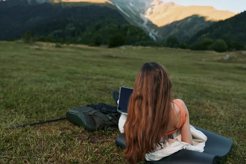 A focused individual sits on a mat in a grassy meadow, using a laptop with scenic mountains in the background, embodying remote work and outdoor serenity.