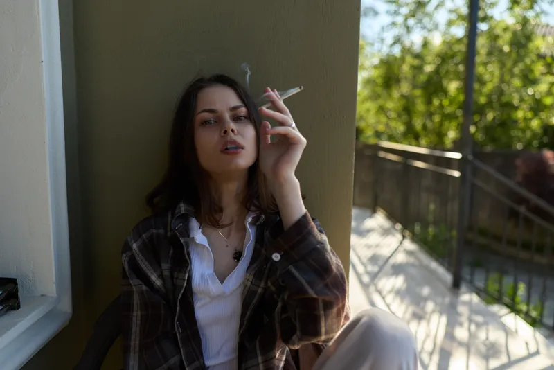 Woman relaxing on balcony wearing casual plaid shirt and white tee, smoking a cigarette, moody urban lifestyle scene with natural light and green trees in background
