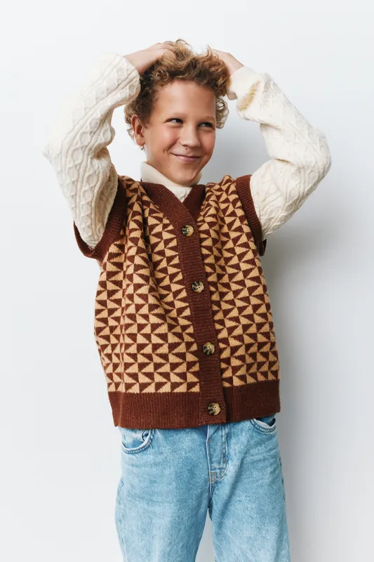 Cheerful young boy poses playfully in a geometric knitted vest and soft cream sweater, creating a warm portrait with casual fashion vibes for family projects.
