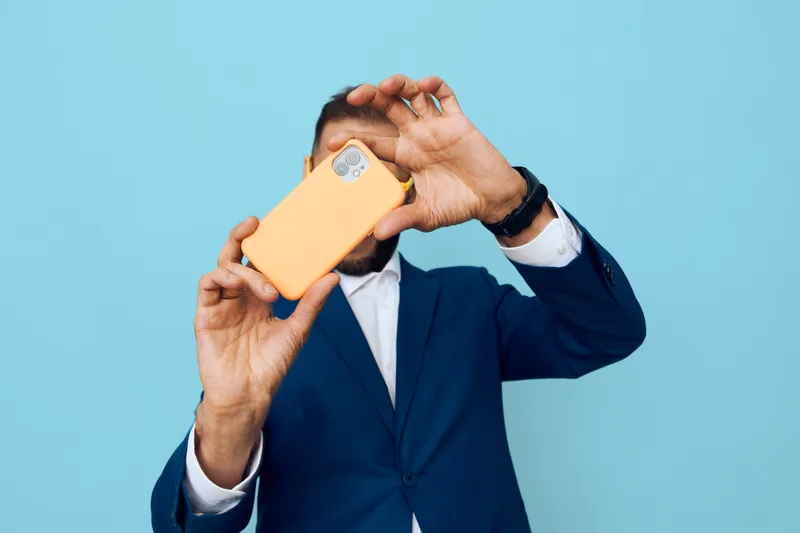 A professional man in a blue suit on a solid colored backdrop, holding an orange phone, posing with confident expression and focused energy to convey success and poise.