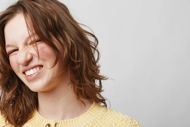 Young woman with curly brown hair wearing yellow sweater smiles with eyes closed on light background. Concept of happiness, joy, natural beauty, and casual lifestyle portrait.