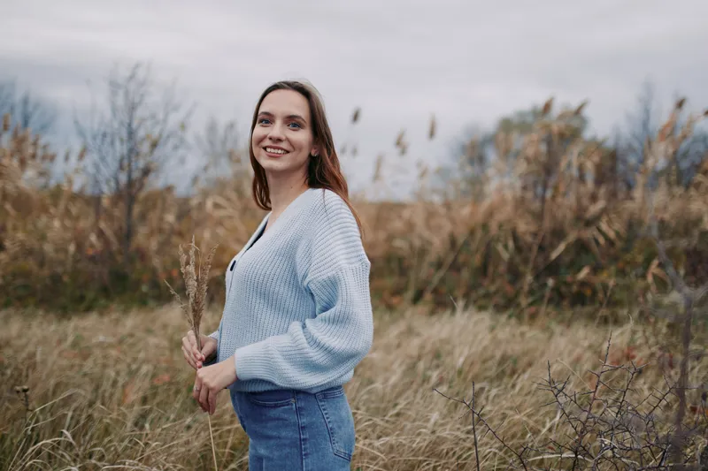 Candid outdoor portrait of a young woman standing in tall grass, smiling naturally and holding dried wildflowers, conveying a genuine, trustworthy mood and honest everyday beauty; soft light enhances