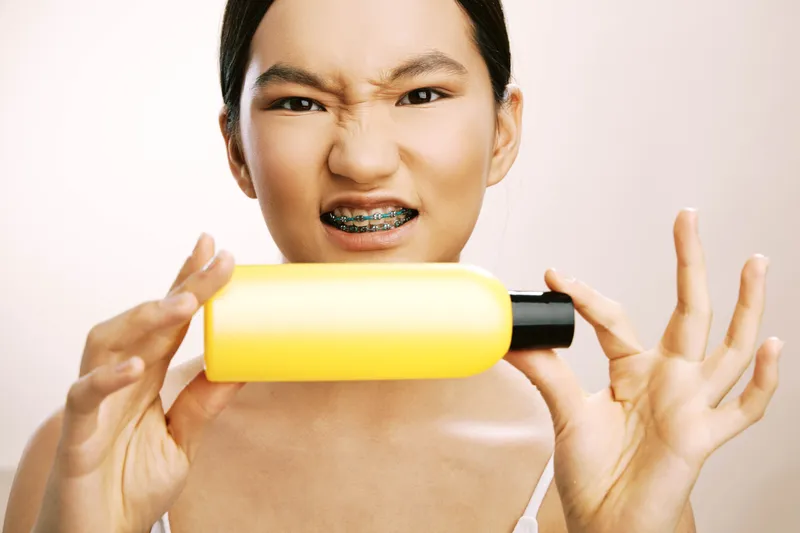 asian woman with braces shows a tense expression while holding a bright bottle in a clean studio, conveying a curious yet uneasy moment as she tests a new product.