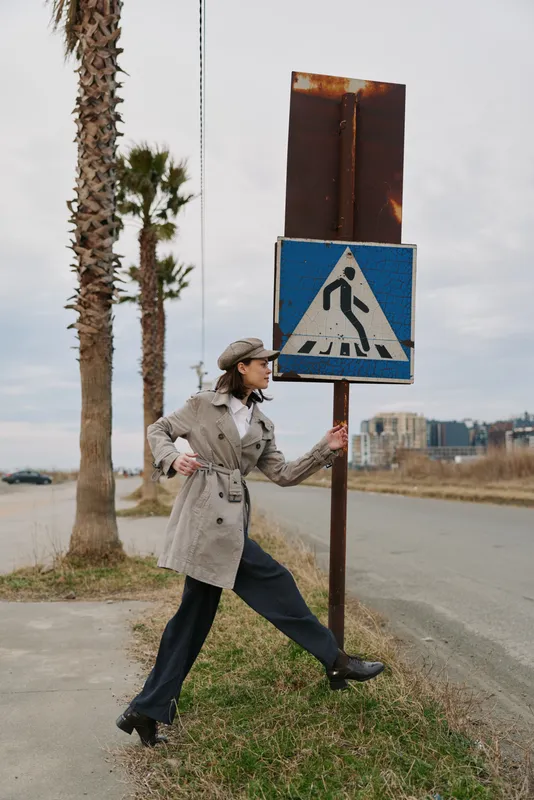 A stylish woman in a trench coat and cap stretches near a pedestrian crossing sign on an empty street with palm trees and overcast sky. Urban fashion and outdoor concept.