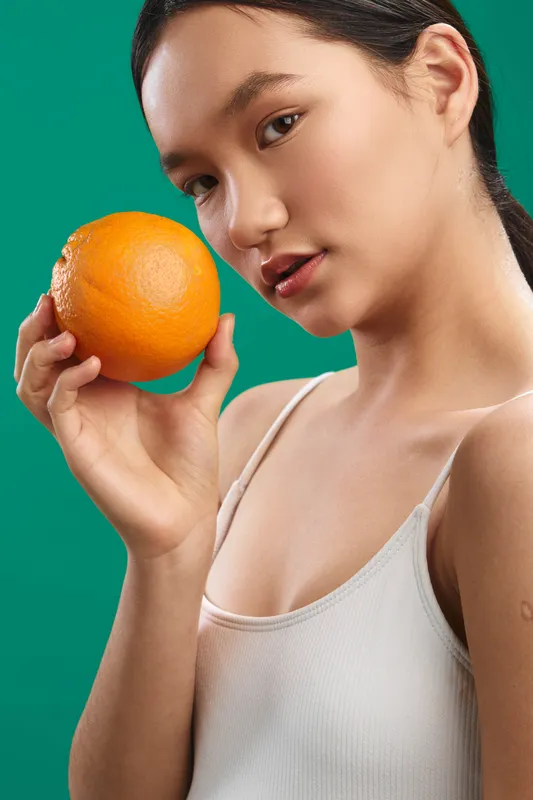 Asian woman holds an orange in a bright studio against a green backdrop, looking at the camera with a calm expression. The vivid color creates a fresh, playful mood suitable for lifestyle and