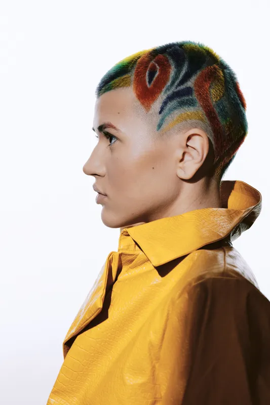 Woman with colorful artistic hair design wearing a yellow coat, showcasing creative fashion, bold hairstyle, and modern style against a white background.
