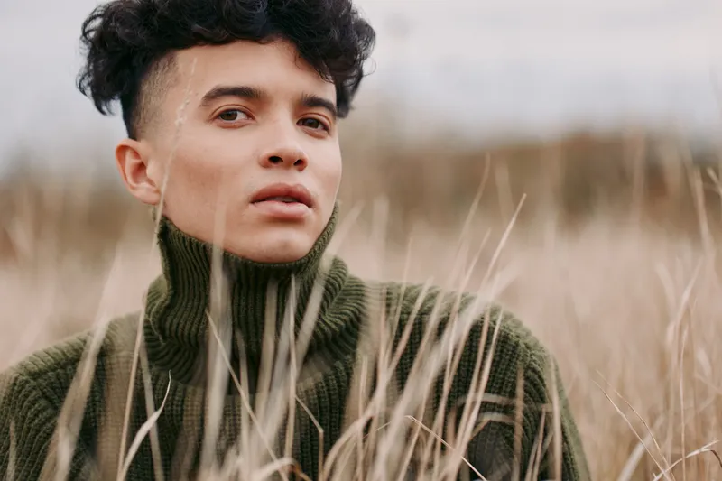 Close candid portrait of a young man in a wool turtleneck amid tall dry grass, conveying authenticity and honest expression with natural texture, realistic skin detail and documentary lifestyle focus.