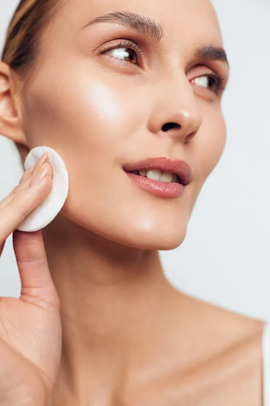 A woman gently wipes her cheek with a cotton pad during a skincare routine, showcasing a radiant complexion, calm focus, and a self care moment in soft lighting.