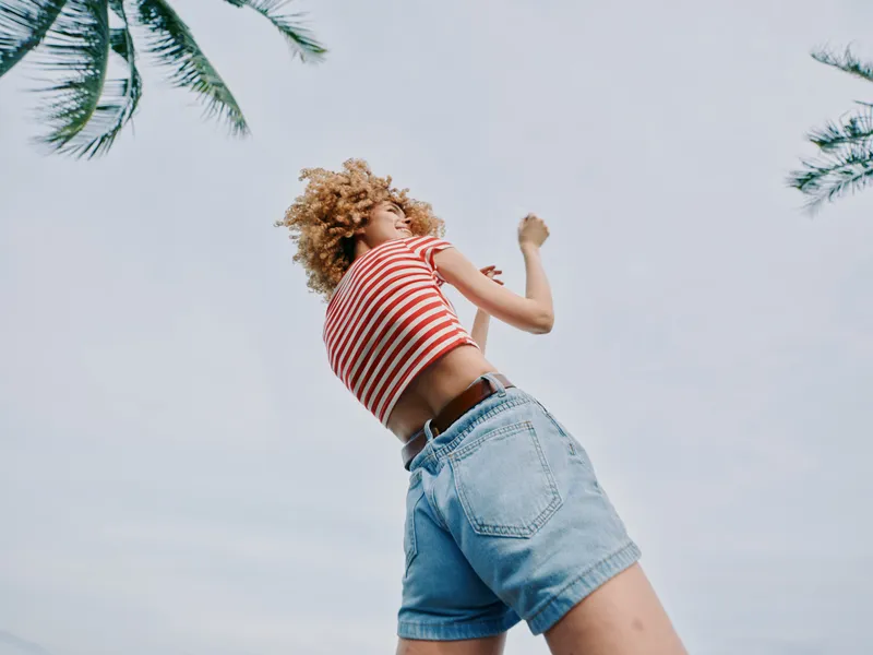 A carefree person leaps into the air, wearing a red and white striped top and light denim shorts, shot from a low angle against a bright sky and palm trees.
