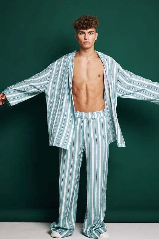 Young man with curly hair wearing striped loose pajamas standing with arms outstretched against dark green background in studio. Casual sleepwear and relaxed pose for fashion or lifestyle.
