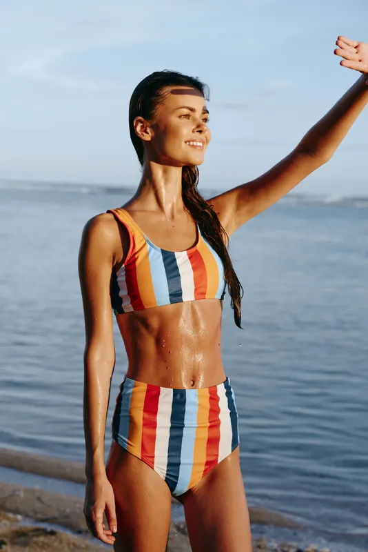 Woman in striped swimsuit stands on beach near sea, smiling in summer sunlight, raised arm pose, youthful vacation portrait, wet skin glow, gentle waves and clear horizon, blue sky