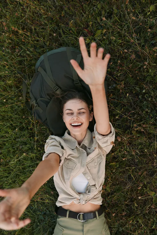 Cheerful woman outdoors, lying on grass, reaching up with one arm, bright smile, sunlit moment, relaxed pose, natural colors, lush greenery background
