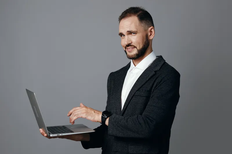 Portrait of a professional man in a suit with a laptop, standing against a solid color background. The scene conveys confidence and focus, showcasing business readiness and calm determination in a