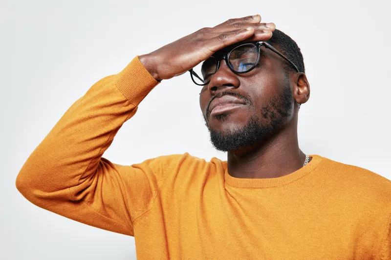 African man with dark skin and beard wearing glasses and orange sweater holding forehead with closed eyes in stress or headache isolated on white background.