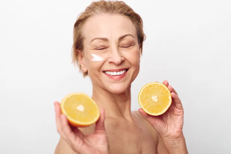 Happy middle aged woman with natural skin care cream on face holds two fresh orange halves smiling with closed eyes on white background. Concept of wellness and healthy lifestyle.