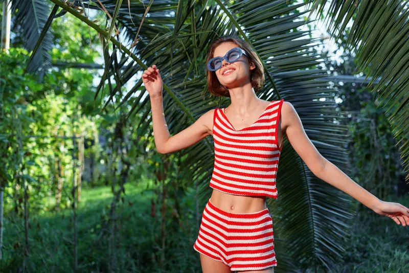 A cheerful woman wearing a red and white striped bikini top and shorts stands outdoors among lush greenery, smiling warmly and enjoying a sunny, carefree moment, exuding bright mood.