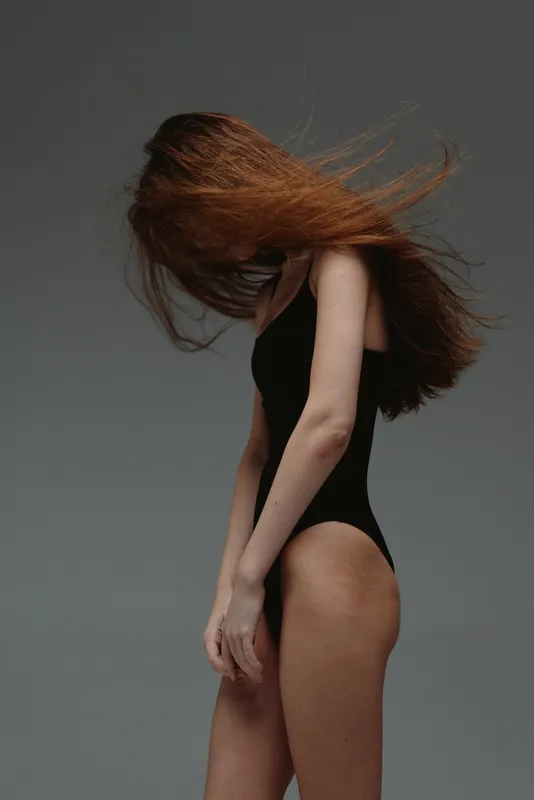 Woman with flowing red hair wearing a black bodysuit standing against a gray background, showcasing natural beauty and movement.