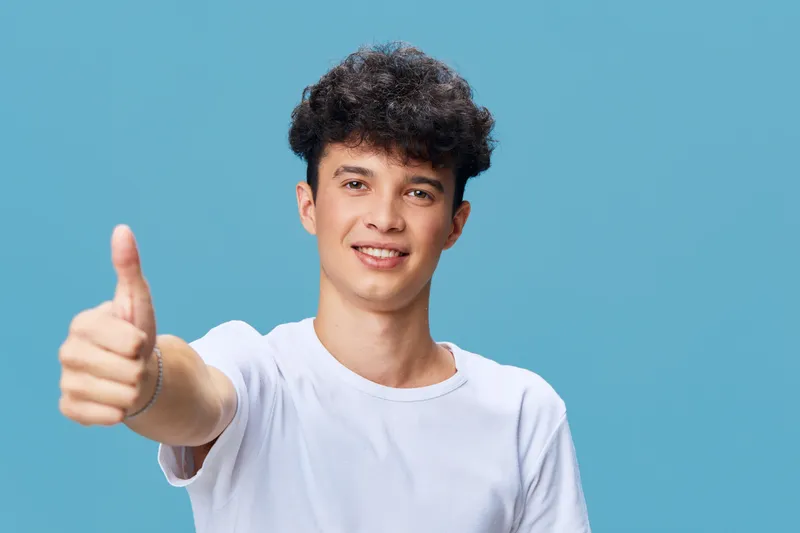 young man showing thumbs up wearing white t-shirt with curly hair on blue background, smiling and expressing positivity, casual style portrait