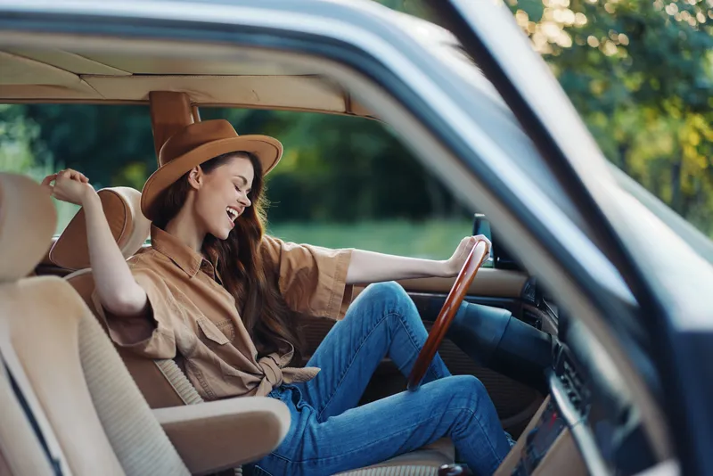 Happy young woman enjoying a road trip in a vintage car, wearing a stylish hat and relaxed outfit, with a natural green background and warm sunlight.