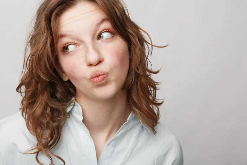 Young woman with curly red hair makes a playful face looking sideways. Casual white shirt and neutral background create a light, fun, and spontaneous mood portrait.