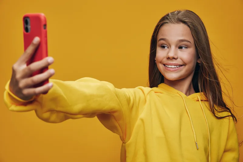 Girl with long hair in yellow sweater taking selfie with smartphone on yellow background smiling and looking aside for social media or communication concept portrait