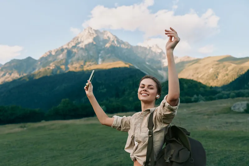 Young woman with a backpack raises her arms in a joyful celebration on a sunny mountain meadow, capturing a candid travel moment amid green hills and distant peaks.