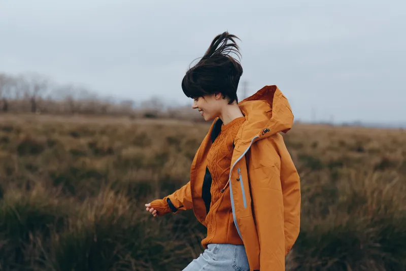 Woman outdoor lifestyle portrait, wearing orange jacket and hood, walking in autumn countryside, windy moment with soft light, relaxed expression, natural colors, blurred fields