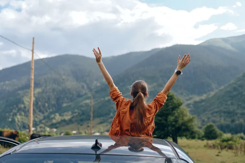 Person with arms raised from an open sunroof enjoying a scenic mountain landscape, conveying freedom, travel spirit, and outdoor adventure