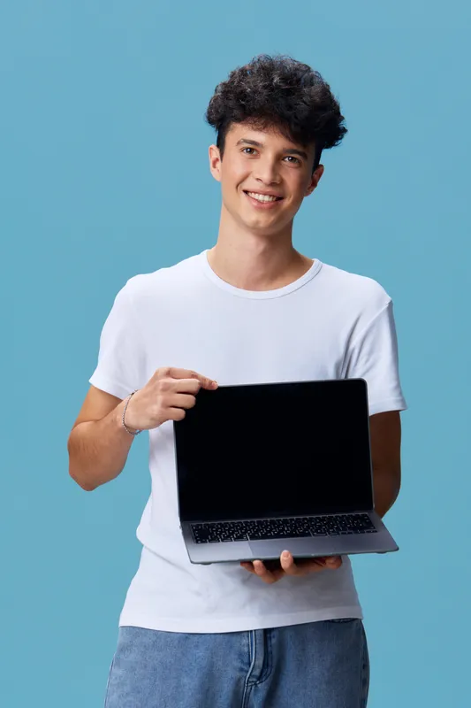 young man holding laptop smiling, casual white t-shirt, curly hair, standing against blue background in bright studio lighting