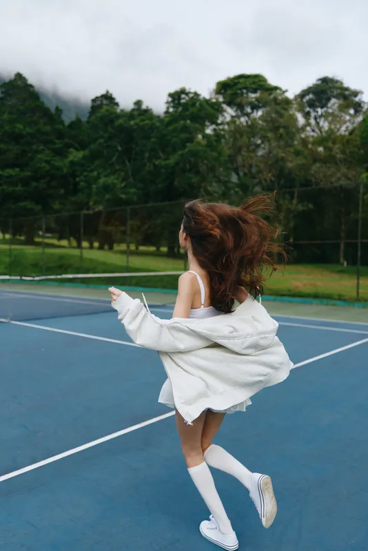 Joyful young woman in sportswear runs on a tennis court amidst lush green nature, wearing a white oversized jacket and knee high socks