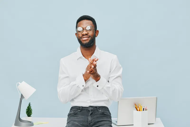 Joyful young man with glasses, wearing white shirt, standing in modern office with cluttered desk and laptop, celebrating success, smiling cheerfully, bright atmosphere, positive vibe, casual style, b
