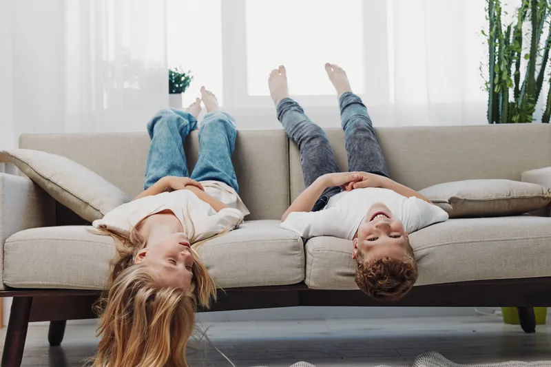Two carefree children lie upside down on a beige sofa in a bright living room with natural light, enjoying playful moments and relaxed weekend fun at home.